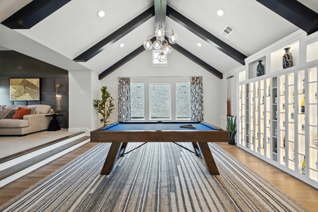 Game room with vaulted ceiling and dark wood beams, a pool table on a striped rug, backlit display shelving, and adjacent cozy lounge area with modern decor.