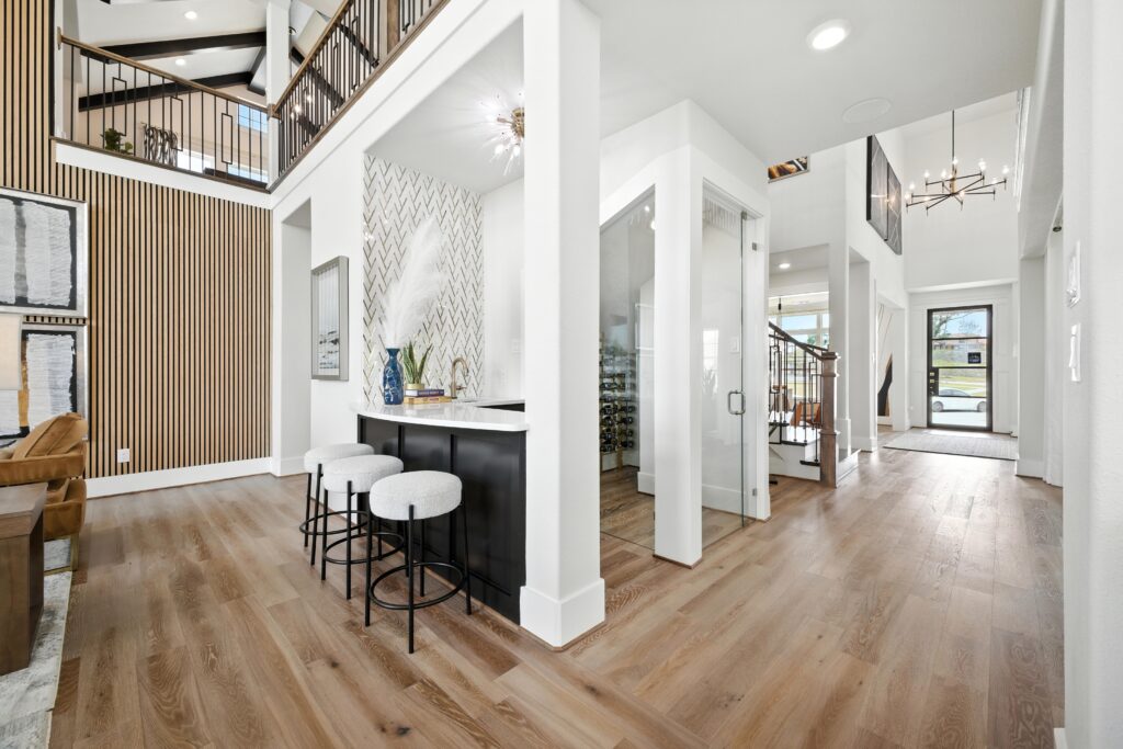 Contemporary open-concept foyer featuring a wet bar with herringbone tile backsplash, natural wood slat accent wall, and a glass-enclosed wine room beside a wooden staircase.
