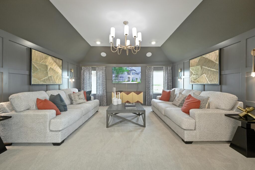 Stylish media room with two large gray sofas, modern chandelier, geometric artwork, paneled dark gray walls, and a wall-mounted TV between curtained windows.